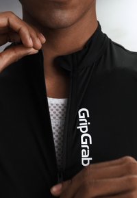 Henkilö pukeutunut mustaan vetoketjulliseen vaatteeseen, jossa lukee "GripGrab", aloittaa sen avaamisen paljastaen valkoisen kuviollisen alusvaatteen.