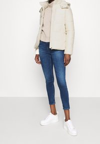 Giacca puffo crema con chiusura a zip, colletto alto e cappuccio, abbinata a un maglione lavorato beige e jeans blu aderenti, completata da sneakers bianche.