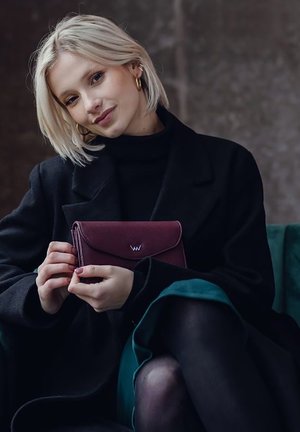Burgundy clutch purse with fold-over design and a silver logo, held by a person wearing a black coat and turtleneck, seated on a green chair.