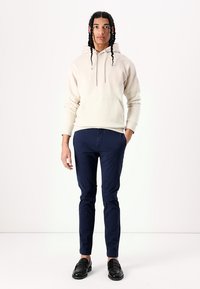 Beige hættebluse med frontlomme, parret med marineblå slim-fit bukser og sorte loafers. Modellen står foran en hvid baggrund.