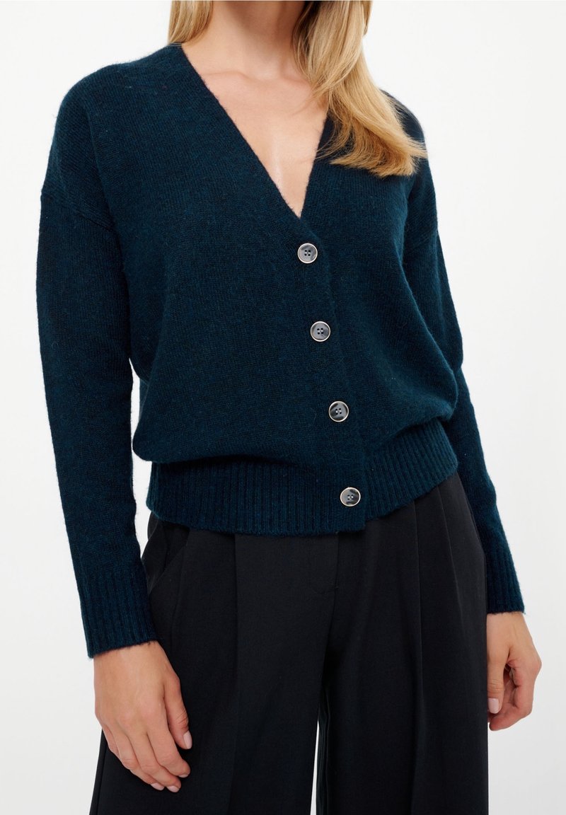 Mörkblå stickad kofta med djup V-ringning och sex silverfärgade knappar. Har ribbade ärmslut och fåll, mjuk textur och avslappnad passform.