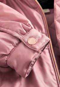 Detail einer rosa gepolsterten Jacke mit einem gerafften Ärmelbündchen, das mit einem roségoldenen Druckknopf und kontrastierender Stepptextur verziert ist.