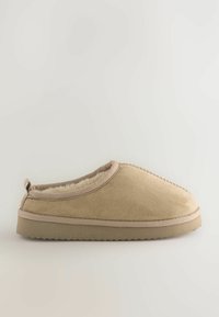 Zapatilla de ante color marrón claro con forro de piel de oveja blanca y suave. Presenta una forma redondeada, diseño de fácil acceso y suela de goma texturizada.