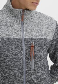 Graue und schwarze gestrickte Fleecejacke mit hohem Kragen, einem Frontreißverschluss und einer Reißverschlusstasche auf der Brust mit einem braunen Kordelakzent.