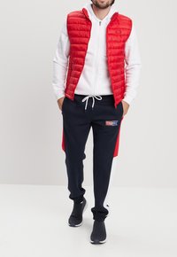 Gilet trapuntato rosso sopra una felpa bianca, abbinato a pantaloni jogger neri con pannelli laterali rossi e bianchi, e scarpe slip-on nere.