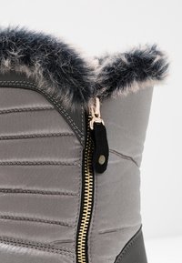 Botín aislante gris con un borde de piel sintética, que cuenta con un zipper dorado y acentos negros. El material es suave, texturizado con detalles acolchados.