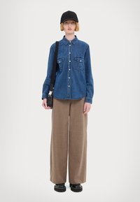 Τζιν πουκάμισο με κουμπιά και δύο τσέπες στο στήθος, συνδυασμένο με φαρδιά καφέ παντελόνια και μαύρες πλατφόρμες, συμπληρωμένο με μαύρο καπέλο.