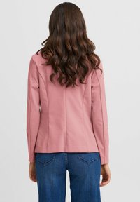 Roze op maat gemaakte jas van een gladde stof, met lange mouwen, een getailleerde pasvorm en achtereen seam details. Gecombineerd met blauwe jeans.