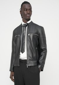 Μαύρο δερμάτινο μπουφάν με ψηλό γιακά, τσέπες με φερμουάρ και υφασμάτινες επιφάνειες με υφή. Φορεμένο με λευκό πουκάμισο και μαύρη γραβάτα. Ραμμένο να φαίνεται κομψό.