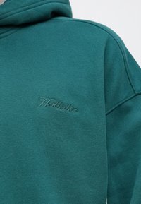 Teal bluza z kapturem wykonana z mieszanki bawełny, charakteryzująca się miękką strukturą, dużym kapturem i haftowanym logo "Hollister" w subtelnej, tonacyjnej odsłonie.