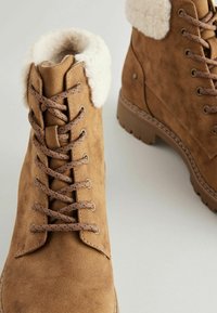 Botas de tobillo de ante marrón con un cuello de piel sintética, puntera redonda, cordones de contraste y suela de goma texturizada. Diseño con cordones y detalle de costura visible.