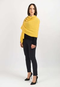 Scialle giallo drappeggiato su una maglietta nera, abbinato a pantaloni neri e tacchi a punta. Lo scialle ha una texture morbida e un design fluido e lineare.