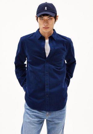 Navy blue corduroy shirt with long sleeves, button closure, and a chest pocket, paired with light blue denim jeans and a navy cap.