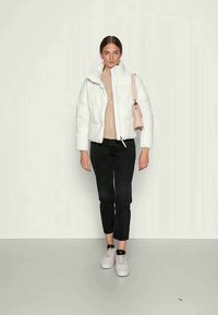 Giacca puffy bianca con colletto alto, abbinata a una maglietta beige e pantaloni cropped neri. Il modello indossa sneakers grigie chiare e tiene in mano una borsa rosa.
