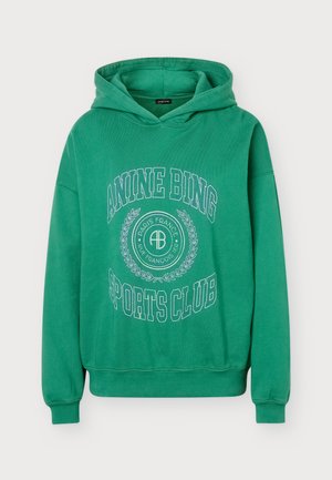 Femme portant un sweat à capuche vert avec le texte "Anine Bing Sports Club", un jean bleu et des chaussures noires, debout devant un fond clair uni.