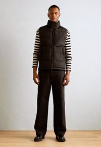 Black puffer vest with high collar over a black-and-white striped long-sleeve shirt, paired with dark trousers and black shoes.
