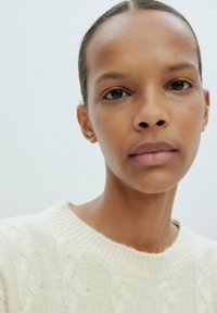 Close-up van een persoon die een lichtcrèmekleurige kabelgebreide trui draagt, met een gladde huid en neutrale make-up, tegen een effen achtergrond.