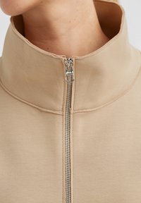 Beige Reißverschlussjacke mit hohem Kragen, glatter Stoffstruktur und metallischem Reißverschlussdetail, das ein minimalistisches Design betont.