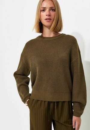 Donna bionda che indossa un maglione verde oliva e pantaloni a righe abbinati, in piedi con una mano in tasca su sfondo bianco.