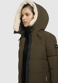Puchowa kurtka w oliwkowym kolorze z dużym beżowym kapturem wyłożonym sztucznym futrem. Posiada czarny suwak i logo marki na rękawie.