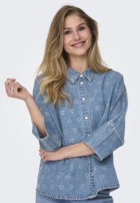 Svetlo modra denim srajca z ovratnikom, sprednjim zapiranjem na gumbe in okrasnimi srčki. Ima tri četrtinske rokave in sproščen kroj.