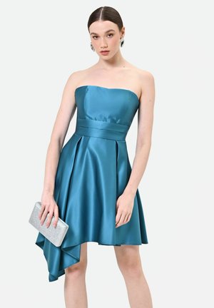 Donna che indossa un vestito di raso turchese senza spalline con un orlo asimmetrico, tiene una clutch argentata e indossa grandi orecchini argentati.