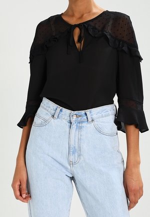 Zwarte doorschijnende blouse met ruches en strikdetail aan de hals, gecombineerd met een lichtblauwe high-waisted jeans, gedragen door een staand persoon.