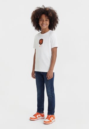 Kind mit lockigem Haar, das ein weißes T-Shirt mit einem orangefarbenen Löwenemblem, blaue Jeans und orange-weiße Nike-Sneaker trägt, lächelt und steht.