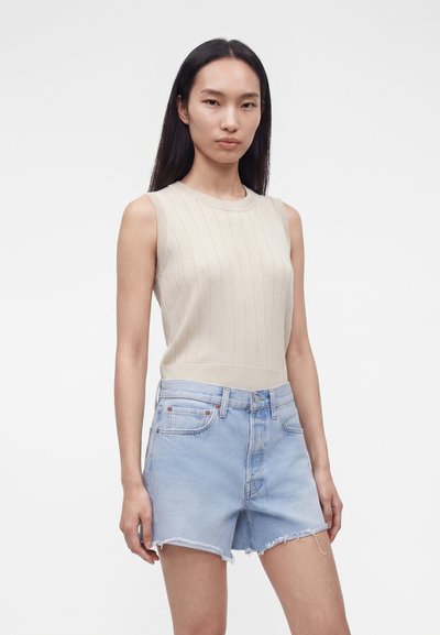 Νέα γυναίκα με μακριά σκούρα μαλλιά, που φοράει μπεζ αμάνικη ριγωτή μπλούζα και ανοιχτό μπλε ψηλόμεσο τζιν σορτς με απλό φόντο.