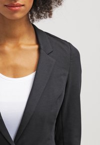 Musta bleiseri, joka on valmistettu kuvioidusta kankaasta, istuu hyvin ja siinä on terävät kaulukset, puettuna valkoisen aluspaidan päälle.