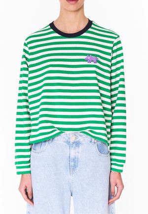 Personne portant un t-shirt à manches longues rayé vert et blanc avec un col noir et un patch animal violet, associé à un jean taille haute bleu clair.