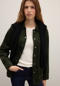 Green corduroy shirt jacket with black sleeveless layering piece, silver snap buttons, and a white sweater underneath. Dark blue jeans visible.