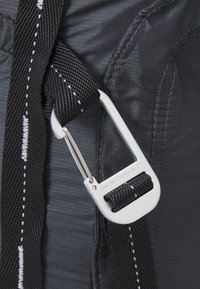 Svart nylonrem med vit sömnad fäst vid en metallkarbinhake. Bakgrunden visar en slät, grå tygyta.