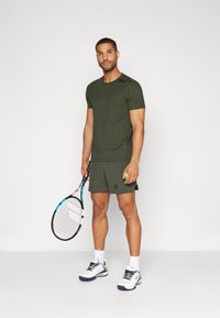 Olive zelena športna majica in kratke hlače z logotipom, drži teniško lopar in žogo. Bele tenis čevlje z modrimi detajli. Enobarvna bela ozadje.