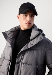 Gray padded jacket with a high collar and zippers, layered over a black turtleneck. Black cap features a square logo patch.