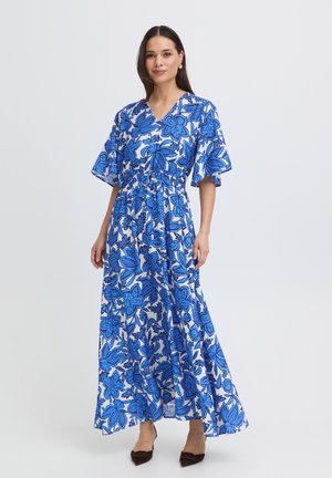 Vrouw draagt een witte maxi-jurk met grote blauwe bloemenpatronen, korte opfladdermouwen en zwarte pumps met spitsneus, staand tegen een effen achtergrond.