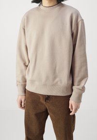 Person som bär en ljusbeige sweatshirt med ribbade ärmslut och nederkant, kombinerat med bruna byxor mot en helt vit bakgrund.