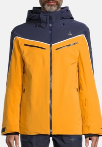 Gelbe und marineblaue wasserdichte Jacke mit Kapuze, ausgestattet mit Reißverschlusstaschen, kontrastierendem Paneeldesign und strukturiertem Stoff.
