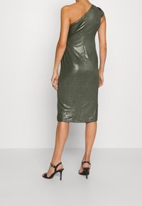 Enaxelad, metallisk grön klänning med draperad design och längd till mitten av vadderna. Buren med svarta, strappar höga klackar.