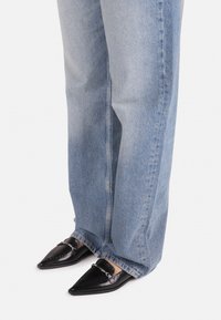 Black patent leather loafers with a pointed toe, decorative chain hardware, worn with light blue baggy denim jeans.