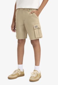 Μπεζ cargo σορτς με πλαϊνή τσέπη, κατασκευασμένα από βαμβάκι, με επίπεδη μπροστινή όψη, θηλιές για ζώνη και λεπτομέρεια με ετικέτα. Συνδυασμένα με μπεζ αθλητικά παπούτσια και λευκές κάλτσες.