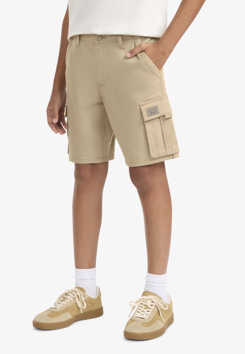 Μπεζ cargo σορτς με πλαϊνή τσέπη, κατασκευασμένα από βαμβάκι, με επίπεδη μπροστινή όψη, θηλιές για ζώνη και λεπτομέρεια με ετικέτα. Συνδυασμένα με μπεζ αθλητικά παπούτσια και λευκές κάλτσες.