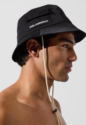 Black bucket hat made from soft, textured fabric, featuring a front patch with "KARL LAGERFELD" in white, adjustable beige drawstring.