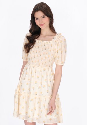 Ung kvinde med langt brunt hår iført en cremefarvet kjole med korte puffede ærmer og gul blomstermønster, smilende let.