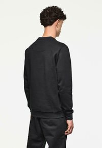 Giovane uomo con capelli mossi e scuri, che indossa una felpa nera a maniche lunghe e pantaloni abbinati, di spalle su uno sfondo bianco.