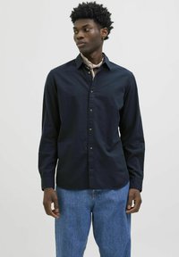 Marineblauwe overhemd met lange mouwen, voorzien van een kraag met knoopsluiting en gouden knopen, gecombineerd met lichtblauwe denim jeans. De stof lijkt glad.