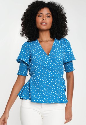 Blaue Bluse mit weißen Punkten, kurzen gepufften Ärmeln mit gerafftem Material und einem Peplum-Saum. Kombiniert mit weißen Hosen.