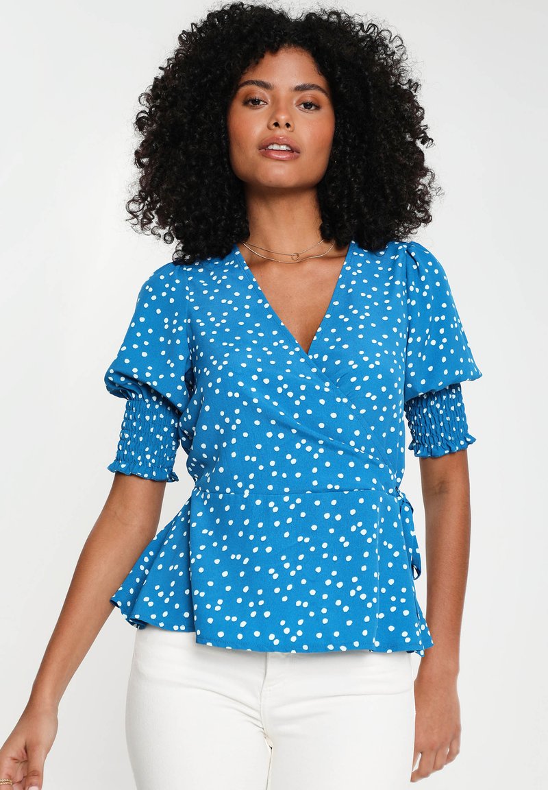 Blaue Bluse mit weißen Punkten, kurzen gepufften Ärmeln mit gerafftem Material und einem Peplum-Saum. Kombiniert mit weißen Hosen.