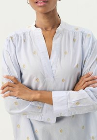 Blusa in cotone azzurro chiaro con scollatura a fessura, ricamo a diamante dorato, maniche lunghe e polsini con bottoni. Tessuto morbido e vestibilità rilassata.