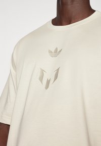 Beige puuvillapaita, jossa on pyöreä kaula-aukko ja hienovarainen kohokuvioitu logo tummahkossa beigen värissä keskellä ja yllä, minimalistinen tyyli.